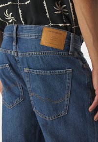 Jeans de denim azul con un parche de cuero y costura en contraste. Presenta dos bolsillos traseros con detalles de costura curva.