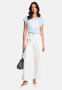 T-shirt bleu clair avec texte blanc, pantalon large taille haute blanc et ballerines blanches pointues. Sac à main à motifs noirs et bleus.