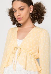 Blusa amarela com padrão floral, de decote em V, cintura franzida e bainha com babados. Tecido leve com mangas longas e bufantes e laços.