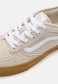 Beige velúr sportcipő fehér fűzővel, kék díszcsíkkal és perforált részletekkel. Rágógumi talp, amely a tapadásra és tartósságra lett tervezve.