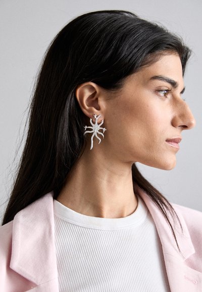 Orecchini a dichiarazione in argento con un design organico e ramificato, caratterizzato da dettagli intricati. Indossati all'orecchio sinistro, in contrasto con capelli scuri.