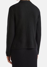 Žena v černé strukturované bundě s dlouhým rukávem a černé žebrované sukni, zobrazená zezadu na bílém pozadí.