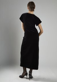 Czarna aksamitna sukienka maxi z krótkimi rękawami, dopasowanym krojem i prostym dołem. Noszona z czarnymi sandałami na wysokim obcasie oraz przezroczystymi rajstopami.