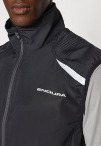 Schwarze Radweste mit hohem Kragen, Vorderreißverschluss, gerippten Schulterakzenten und einem reflektierenden weißen Streifen an der Seite. Markenlogo "ENDURA" abgebildet.