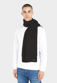 Sciarpa nera a coste in maglia con una texture morbida e un'etichetta firmata, avvolta attorno al collo di una figura che indossa un maglione bianco e jeans blu.