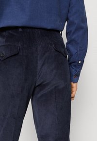 Navy corduroy trousers with ribbed texture, featuring back pockets and black buttons. Paired with a blue long-sleeve shirt.