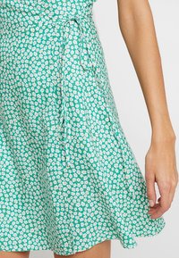 Green floral dress with a wrap design, featuring small white flowers on a textured fabric, and a tie at the waist for adjustable fit.