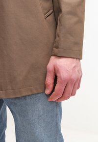 Gros plan d'une main reposant sur le côté, portant une veste marron et un jean en denim bleu clair sur un fond uni.