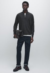 Gray zip-up sweater made of textured fabric, layered over a white shirt. Dark denim jeans, brown leather shoes, and holding a gray jacket.