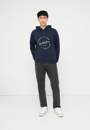 Mladi moški, oblečen v temno modro jopo Jack & Jones, temno sive kavbojke in bele superge, stoji pred enobarvnim belim ozadjem.