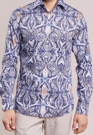 Mann trägt ein langärmliges, geknöpftes Hemd mit aufwendigen blauen, weißen und beigen Paisley- und Blumenmustern, kombiniert mit hellbeigen Hosen.