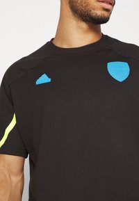 Černé sportovní tričko s krátkým rukávem. Obsahuje modré logo Adidas a znak Arsenalu. Neonově žlutý akcent na rukávu. Hladká textilie.