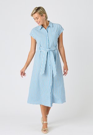 Mujer caminando que lleva un vestido abotonado con cuadros azules y blancos, con cinturón en la cintura y sandalias beige de tacón sobre un fondo liso.