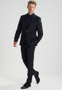 Marineblå dress med en tettsittende blazer og bukser med flat front, laget av glatt stoff. Kombinert med en matchende skjorte og svarte sko.