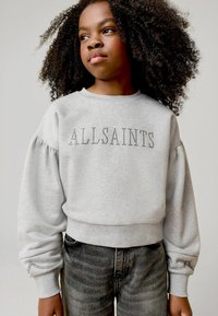 Siva dukserica s puf rukavima, sa natpisom "ALLSAINTS" od srebrnih zakovica. Nosi se uz sive traperice, prikazujući ležeran, teksturiran izgled.