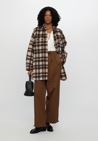 Chaqueta de cuadros marrón y beige sobre una blusa blanca, combinada con pantalones anchos marrones y zapatos negros, sosteniendo un pequeño bolso negro.