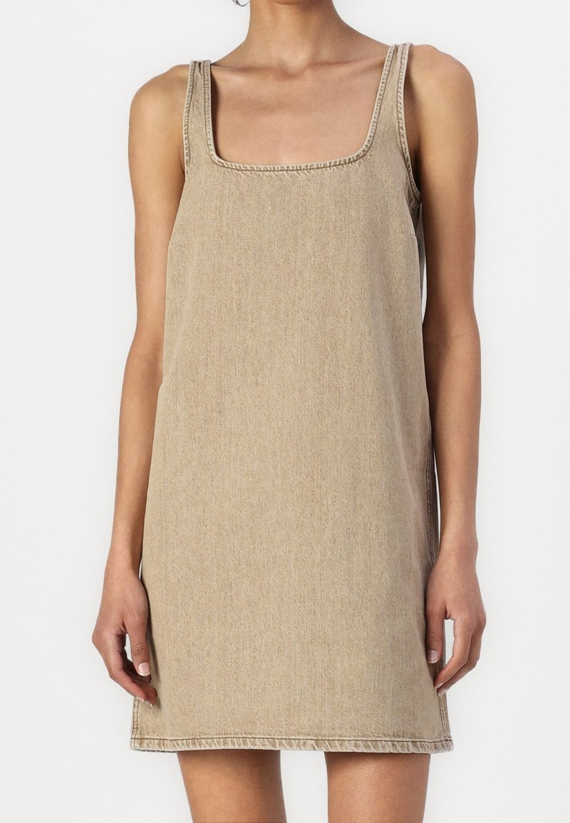 Mini-robe sans manches en denim beige avec un décolleté carré portée par une personne debout sur un fond uni.