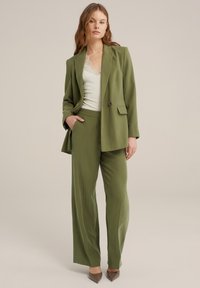 Blazer entallado verde oliva y pantalones de pierna ancha de tejido suave. El blazer presenta un botón único y bolsillos, combinado con una blusa blanca con encaje.
