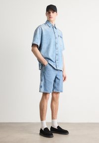 Camisa de mezclilla de color azul claro con mangas cortas, combinada con pantalones cortos de mezclilla a juego. Una gorra negra, mocasines negros y calcetines blancos completan el conjunto.