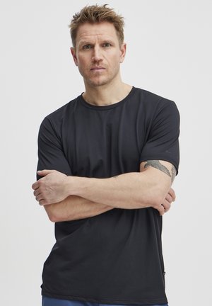 Camiseta deportiva negra hecha de tela suave. Presenta un cuello redondo, mangas cortas y un logotipo sutil en la manga. Diseño simple.