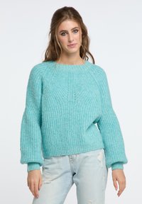 Maglione turchese lavorato a maglia con un motivo testurizzato, scollatura rotonda e maniche leggermente a sbuffo. Abbinato a jeans in denim chiaro con effetti strappati.