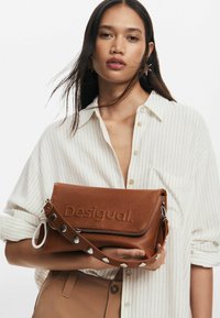 Borsa a tracolla in pelle marrone con logo "Desigual" impresso, chiusura con zip e una tracolla removibile con borchie in tono argentato. Design rettangolare, texture liscia.