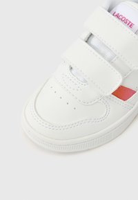 Λευκό αθλητικό παπούτσι με ιμάντες με κλείσιμο χιαστί, λείο δερμάτινο υλικό, αναπνεύσιμη επένδυση, ροζ λεπτομέρεια και οπές στο άκρο του δακτύλου.