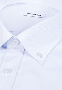 Chemise blanche en tissu texturé, col boutonné et boutons transparents, avec une étiquette de marque "seidensticker" à l'intérieur du col.