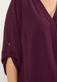 Bluse in Pflaumenfarbe mit lockerer Passform, V-Ausschnitt, hochkrempelbaren Ärmeln, die mit einem Knopf fixiert sind, und zwei Fronttaschen. Glattes, strukturiertes Material.