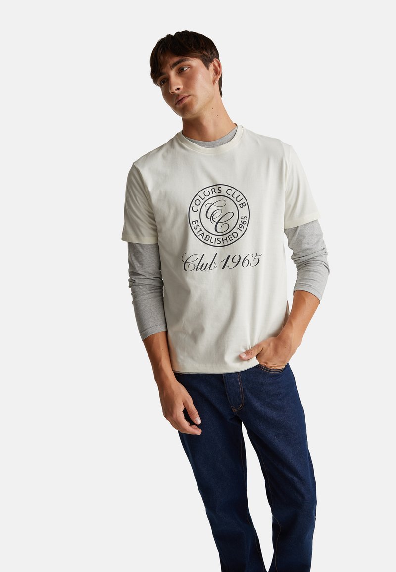 Světle šedé tričko s kulatým černým logem a textem, přes které je navlečeno šedé tričko s dlouhým rukávem. Kombinováno s tmavě modrými džíny, což vytváří ležérní vzhled.