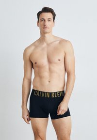 Schwarze Boxershorts mit einem breiten goldenen Bund, auf dem "CALVIN KLEIN" in fetten Buchstaben steht. Glatter Stoff mit einer engen Passform.