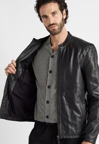 Zwarte leren jas met gewatteerde schouderaccenten en een ritssluiting aan de voorkant, gedragen over een grijs en zwart gestreept overhemd met knopen.