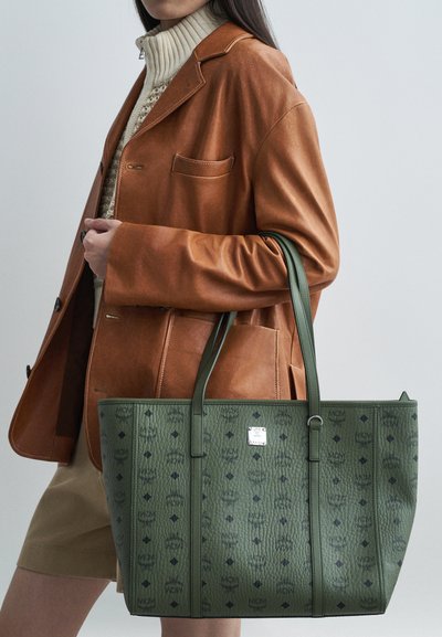 Moteris, vilkinti rudą odinį švarką ir smėlio spalvos megztinį, ant rankos laiko didelę žalią MCM rankinę su raštu.