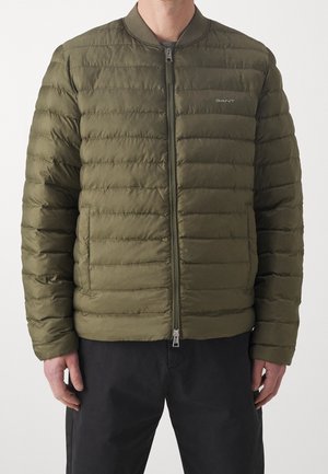 Man wearing an olive green quilted jacket with front zipper and side pockets, paired with black pants, standing against a plain background.