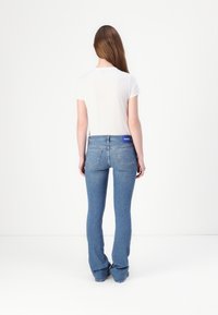 Jean évasé bleu clair avec une taille mi-haute, cinq poches et une étiquette de marque visible à l'arrière. Associé à un top blanc à manches courtes.