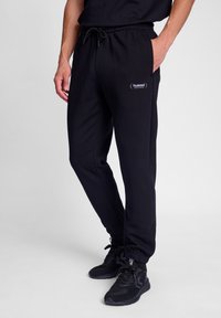 Schwarze Jogginghose mit elastischem Bund und verstellbarem Kordelzug. Sie verfügt über Seitentaschen und ein kleines Logo am linken Oberschenkel.