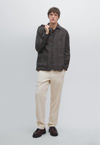Young man standing against plain background wearing a brown checkered jacket, cream pants, dark shoes, holding a folded umbrella over his shoulder.