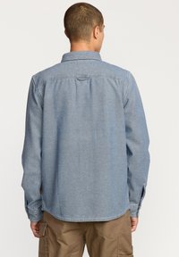 Camisa de mezclilla azul claro con mangas largas, un collar y puños abotonados. Presenta un canesú en la parte posterior y un dobladillo redondeado.