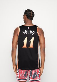 Camiseta de baloncesto negra con "YOUNG" y el número "11" en un degradado naranja. Presenta el logo de la NBA y un contraste de ribete naranja en los hombros.