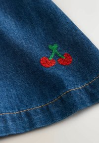 Denimstof med røde broderede kirsebær med en grøn stilk på en blå baggrund, suppleret af en synlig gylden trådsy. 