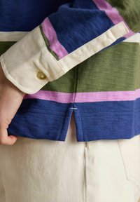 Tuvplānā cilvēka fotogrāfija, kurš valkā daudzkrāsainu svītrains krekli ar krēmbaltiem piedurknēm un gaiši bēšām biksēm, roka atpūšas netālu no apakšmalas.