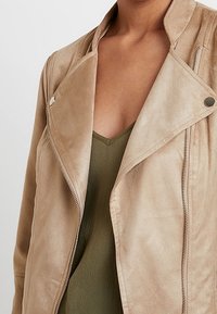 Béžová semišová bunda s klopovým límcem, asymetrickým zapínáním na zip a hladkou texturou. Nositelná přes zelený tílkový top s výstřihem do V.