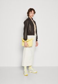 Bruine leren bomberjack, beige top, witte midi-rok, gele gekruiste clutch en grijs-gele sneakers met textuurdetails.