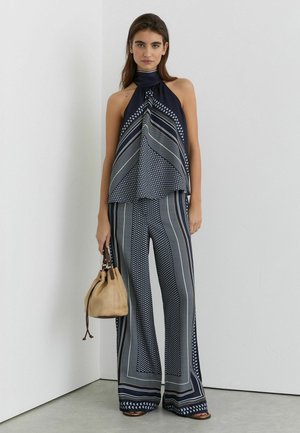 Noor naine kannab tumesinist mustrilist kaelusega topi ja sobivaid laia säärega pükse, hoides käes beežikat paelaga käekotti, seistes valgete seinte taustal.