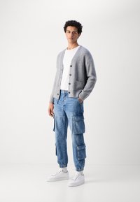 Cárdigan de punto gris con botones de madera, llevado sobre una camiseta blanca. Combinado con jeans cargo azul claro y zapatillas blancas.