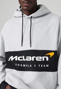 Szara bluza z kapturem z czarnym pasem, na którym widnieje napis "McLaren" oraz "Formula 1 Team" w białym kolorze, akcentowana pomarańczowym logo.