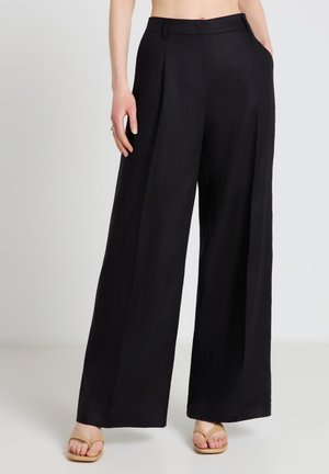 Donna che indossa pantaloni neri a vita alta, a gamba larga, con tasche laterali, abbinati a sandali infradito beige, in piedi su uno sfondo semplice.