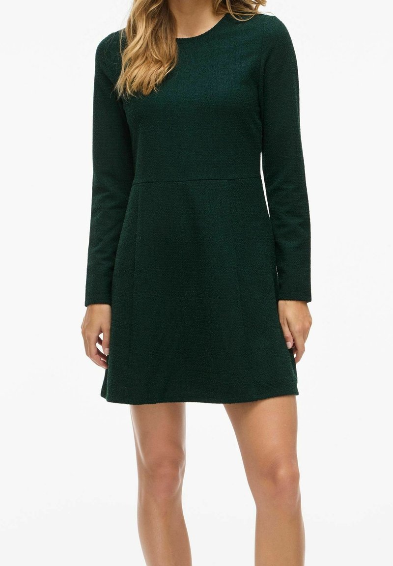 Femme portant une robe verte foncée à manches longues et longueur genou avec un col rond, debout devant un fond blanc uni.