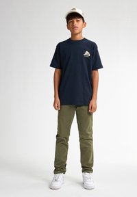 Marineblauwe katoenen t-shirt met een wit logo op de borst, gecombineerd met groene katoenen broek en witte sneakers. Een eenvoudige pet maakt de outfit compleet.