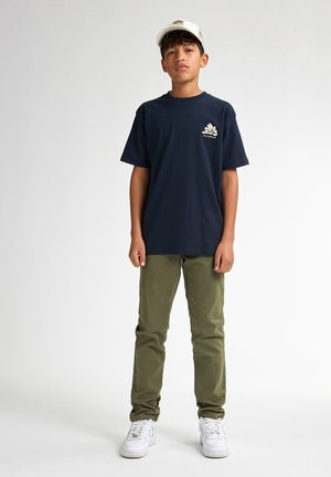 Marineblauwe katoenen t-shirt met een wit logo op de borst, gecombineerd met groene katoenen broek en witte sneakers. Een eenvoudige pet maakt de outfit compleet.